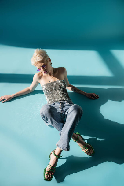 overhead view of tattooed albino woman with heeled sandals and jeans posing on blue  - Fotografie, Obrázek