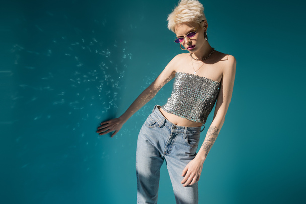 tattooed albino model in trendy sunglasses and stylish outfit posing near blue wall  - 写真・画像