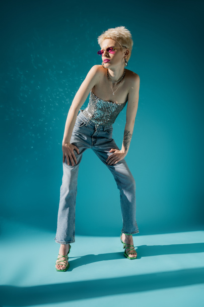 full length of tattooed albino model in trendy pink sunglasses and fashionable outfit posing on blue  - Foto, afbeelding