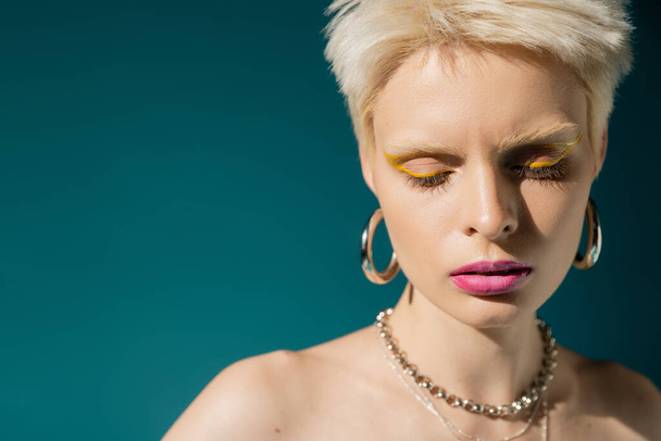 portrait of blonde albino woman with trendy makeup posing in jewelry on blue background  - 写真・画像