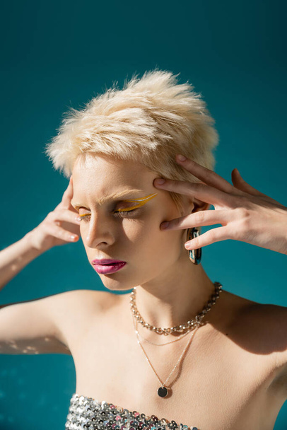 portrait of blonde albino woman with trendy makeup posing with hands near head on blue  - Zdjęcie, obraz