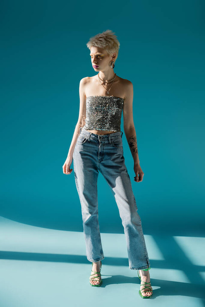 full length view of tattooed albino model in top with sequins and denim jeans standing on blue  - Фото, изображение
