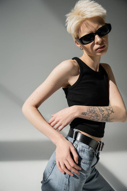stylish blonde woman with tattoo posing in black sunglasses on grey  - Foto, imagen