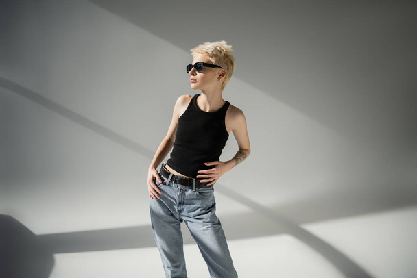 blondynka i wytatuowany model w czarnych okularach przeciwsłonecznych pozowanie na szarym tle  - Zdjęcie, obraz