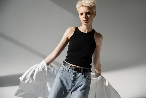 tattooed albino woman in black tank top and jeans taking off white shirt on grey background - Φωτογραφία, εικόνα