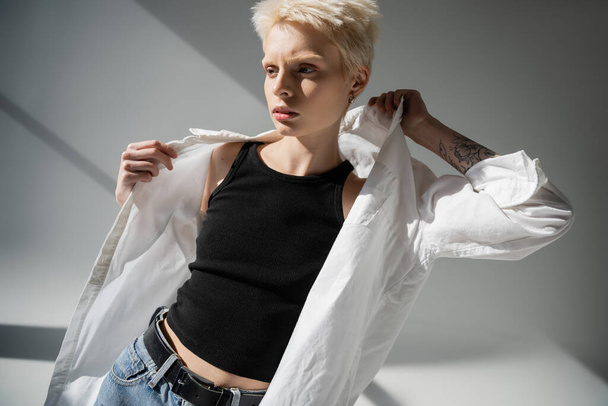 pretty blonde woman with tattoo on hand wearing white shirt on grey background with shadows - Photo, Image