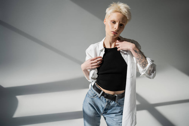 young albino woman with tattoo on hand wearing white shirt on grey background with shadows - Photo, image