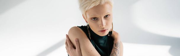 młoda albinoska z tatuażem na dłoni przytulająca się na szarym tle z cieniami, sztandar  - Zdjęcie, obraz