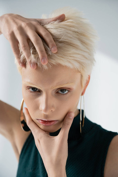 young albino model with fair-skin touching face with hands while posing on grey  - Фото, изображение
