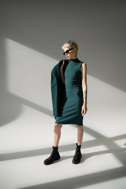 full length of tattooed and blonde woman in sunglasses posing in green dress and coat on grey background  - Foto, Bild