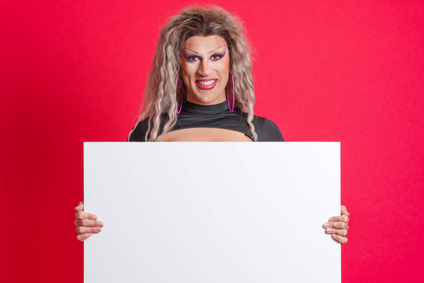 Smiley transgender persona con make up in possesso di un pannello bianco in studio con sfondo rosso - Foto, immagini