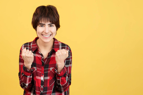 Androgynous person celebrating while raising the fists in studio with yellow background - Photo, Image