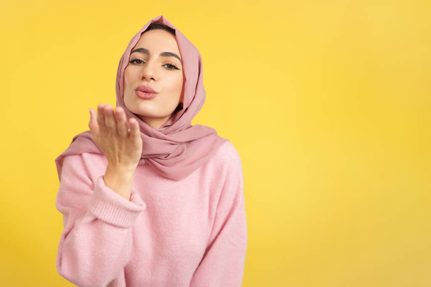 Muslimská žena foukání polibek při pohledu na kameru ve studiu se žlutým pozadím - Fotografie, Obrázek