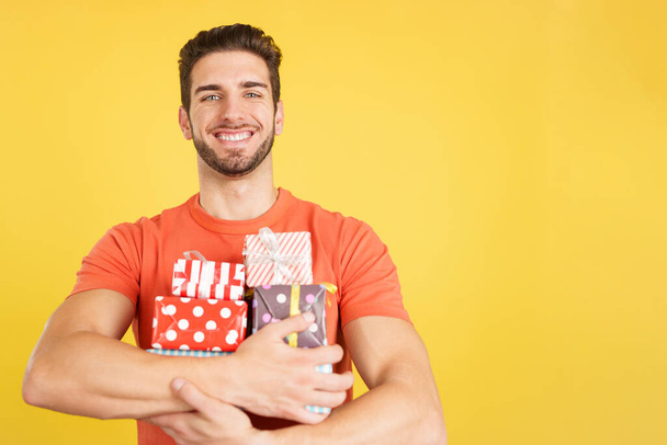Happy caucasian man holing many colorful gifts in studio with yellow background - Φωτογραφία, εικόνα