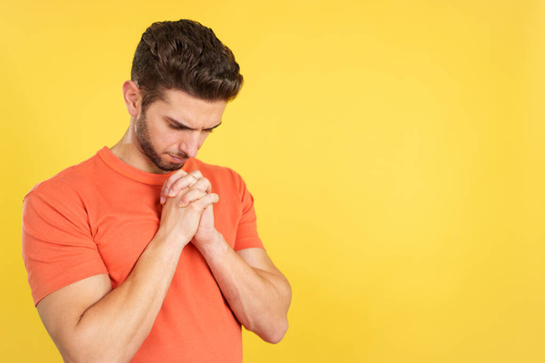 Caucasian man praying with folded hands looking down in studio with yellow background - Foto, imagen