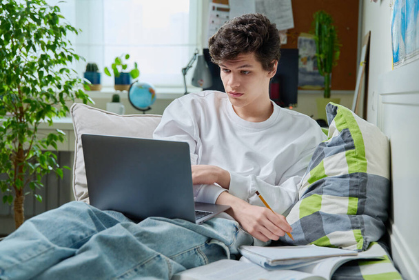 Teenage guy lying at home on couch student using laptop notebook textbook. Handsome male teenager college, high school student learning online remotely. Education, learning, technology, youth concept - Photo, Image