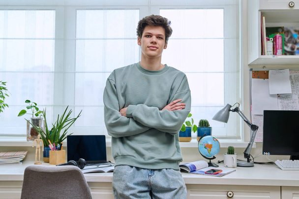 Portrait of handsome guy 18, 19 years old looking at camera in home interior. Confident young male with crossed arms, near window. Lifestyle, youth concept - 写真・画像