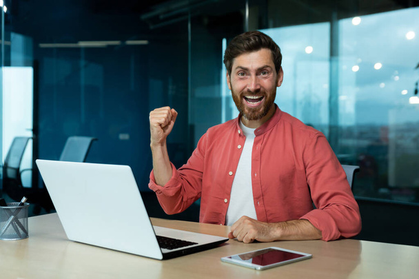 Happy young student sitting in office, campus in front of .aptop, showing victory success gesture with hand, celebrating, looking at camera, smiling. - Fotografie, Obrázek