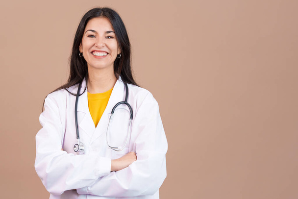 Femme médecin caucasienne souriant à la caméra avec les bras croisés en studio avec fond brun - Photo, image