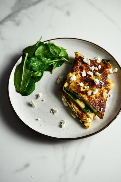 Folded omelette stuffed with spinach and feta on the white plate with spinach leaves on back. Healthy and dietary breakfast dish. Minimalistic food photo. White marble background. . High quality photo - Fotografie, Obrázek