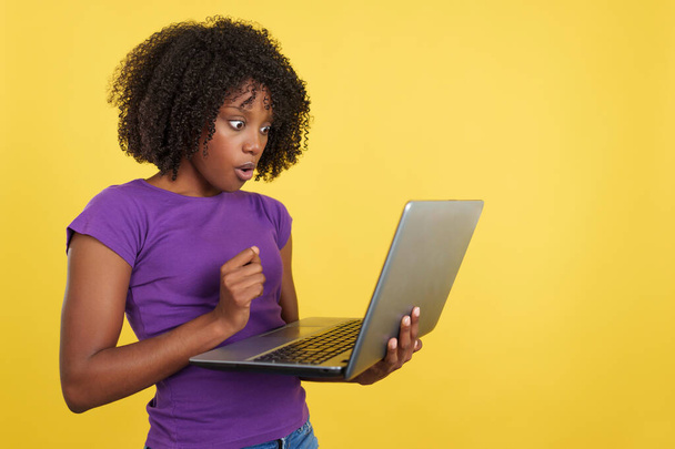 Woman with afro hair looking a laptop with shocked expression in studio with yellow background - 写真・画像