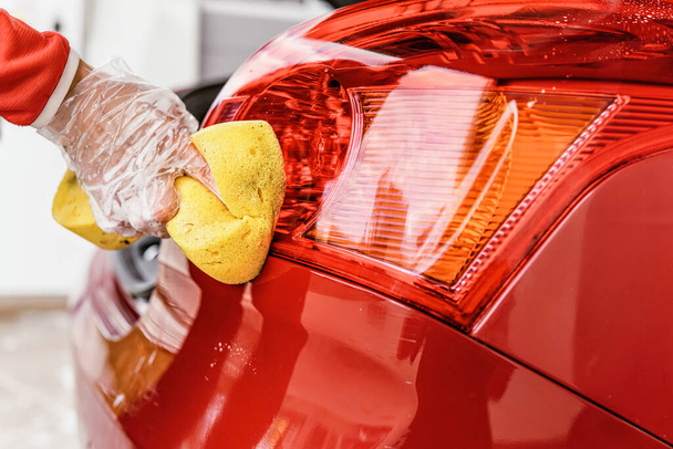 Young woman cleaning rear lights of her car, closeup detail on hand in glove holding yellow sponge - Φωτογραφία, εικόνα