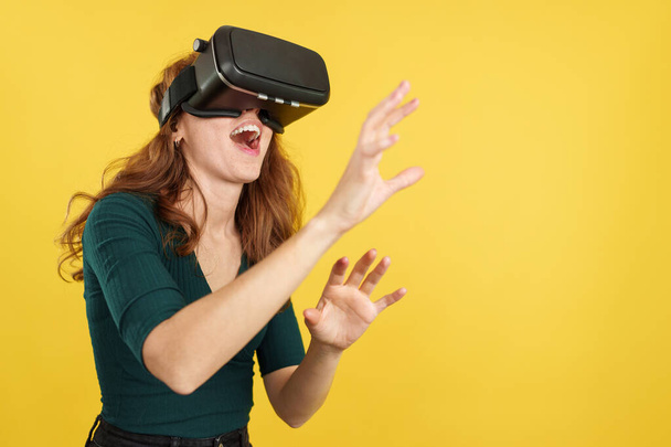 Happy redheaded woman with Virtual Reality goggles in studio with yellow background - Photo, Image