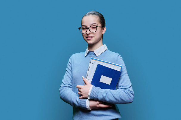 Portrait of smiling high school student girl with textbooks notebooks in hands on blue studio background. Smart friendly teenage female looking at camera. Adolescence, education, study concept - Photo, image
