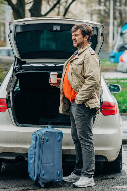 Senior man with cup of coffee and travel bag stands next to car, Wroclaw, Πολωνία - Φωτογραφία, εικόνα