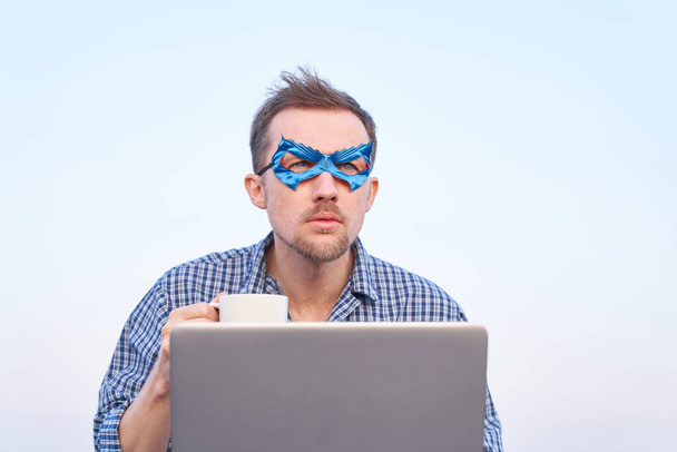 Homem alegre em camisa de camisola e máscara facial azul usando laptop ao ar livre. Suporte de TI cara super-herói usando laptop segurando xícara de chá quente ou café. Imagem de alta qualidade - Foto, Imagem