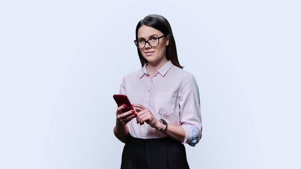 Mujer joven posando con teléfono inteligente en sus manos sobre fondo blanco del estudio. Hermosa mujer de 30 años con gafas de camisa mirando a la cámara. Estudio, carrera, trabajo, estilo de vida, concepto de ocio - Foto, Imagen