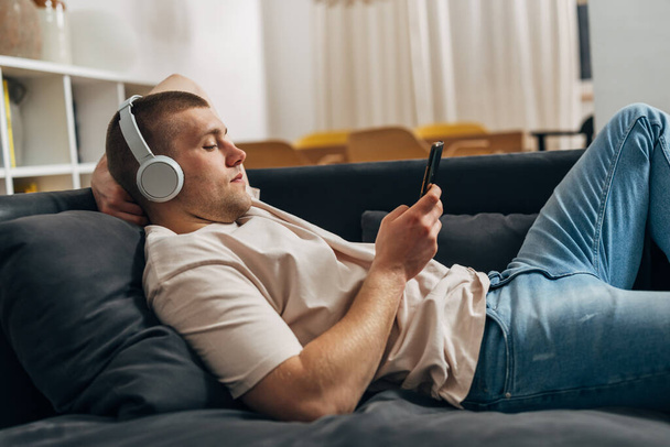 Side view of a Caucasian man resting on the sofa with his smartphone - Photo, Image