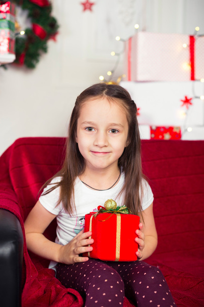 Little girl sitting on couch holding red box, gift, Christmas tree in the background - Fotografie, Obrázek