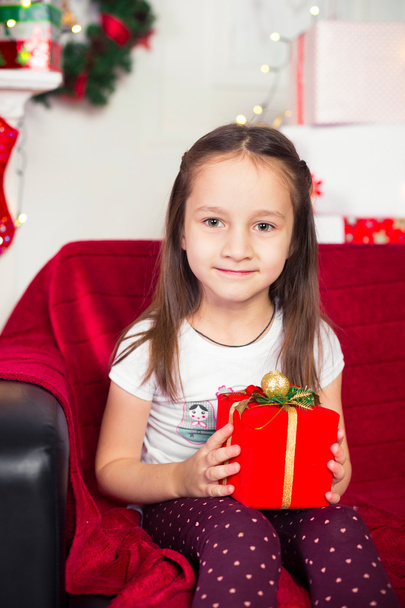 Little girl sitting on couch holding red box, gift, Christmas tree in the background - 写真・画像