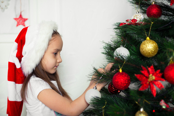 Kleines Mädchen mit roter Mütze hängt am Weihnachtsbaum - Foto, Bild