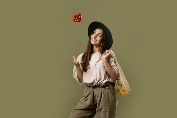 Vegetarian woman wear casual clothes and black hat hold net eco bag with vegetables and throw up a red pepper in air - Фото, изображение