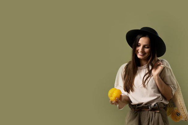 Eco friendly smiling woman in beige t-shirt and black hat throw up a yellow pepper in a air by one hand - Fotografie, Obrázek