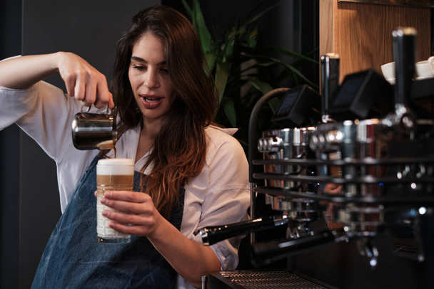 A happy barista preparing fresh coffee and adding milk into a glass while - Фото, зображення