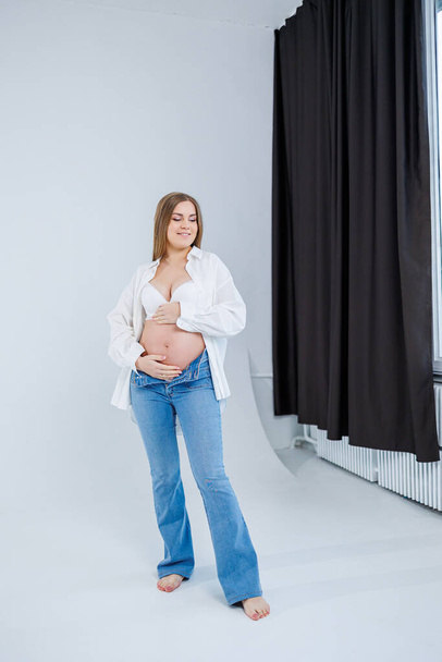 Μια τρυφερή νεαρή έγκυος γυναίκα με λευκό πουκάμισο και τζιν αγκαλιάζει την κοιλιά της. Έγκυος ευτυχισμένη γυναίκα σε λευκό φόντο. - Φωτογραφία, εικόνα