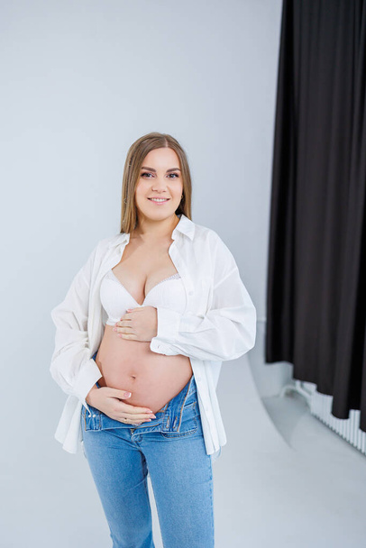 A tender young pregnant woman in a white shirt and jeans hugs her belly. Pregnant happy woman on a white background. - Fotó, kép