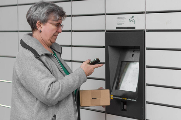 Pniewy, Poland - 22 March 2023. Senior woman with smartphone entering code to open shopping locker in inPost delivery service for receives or sends a box. E-commerce shipping and delivery concept. - Photo, Image