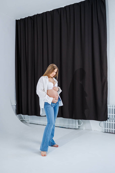 Een tedere jonge zwangere vrouw in een wit shirt en jeans knuffelt haar buik. Zwangere gelukkige vrouw op een witte achtergrond. - Foto, afbeelding