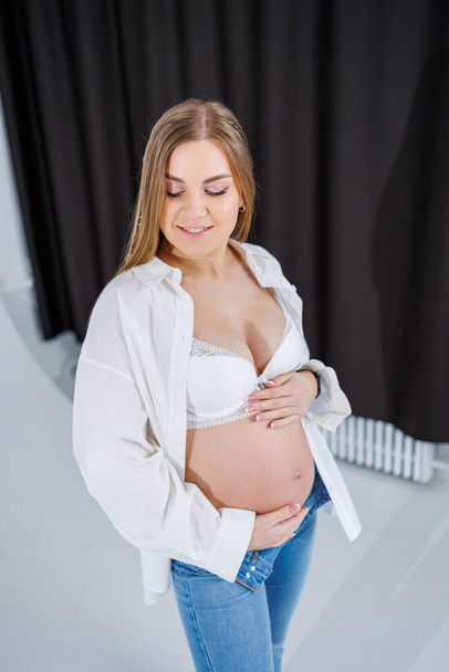 A tender young pregnant woman in a white shirt and jeans hugs her belly. Pregnant happy woman on a white background. - Foto, Imagem