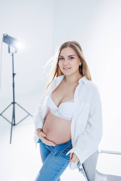 Cute pregnant woman in bra and white shirt on white background. Happy pregnant woman stroking her belly. - Фото, изображение