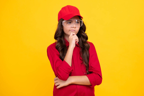 Worker teenager child wearing overalls red, cap and protect glasses. Studio shot portrait isolated on yellow background. Pointing and showing concept. Thinking pensive clever teenager girl - Foto, imagen
