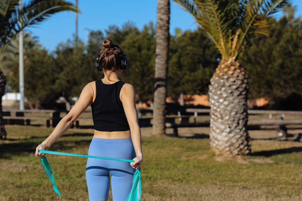 Rückenansicht einer jungen athletischen Frau in Sportbekleidung, die an Sommertagen im Park mit Palmen Übungen zur Stärkung des Rückens und der Arme mit Fitness-Gummiband macht. Weltgesundheitstag - Foto, Bild