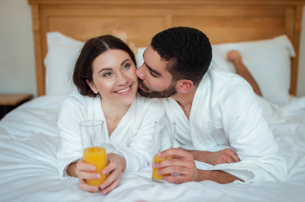 Honeymoon. Loving Boyfriend Kissing Girlfriend Holding Orange Juice In The Morning, Lying Relaxing In Bed In Hotel Suite Indoors. Multiethnic Spouses Enjoying Romantic Vacation - Photo, Image