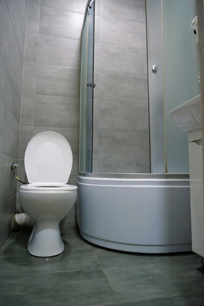 Toilet room interior with white toilet bowl with open lid, shower cabin, wash basin, hairdryer, cabinet under the washbasin and grey wall and floor tiles - Φωτογραφία, εικόνα
