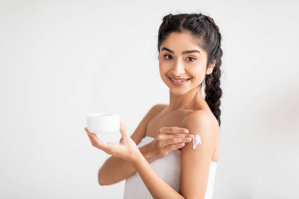 Self Pampering Concept. Beautiful Indian Lady Applying Body Cream On Shoulder, Happy Eastern Woman Wrapped In Towel Using Moisturising Skin Lotion After Bath Standing On White Background, Copy Space - Zdjęcie, obraz