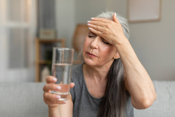 Unhappy sad caucasian old lady hold glass of water and touching forehead, suffering from fever, menopause in living room interior. Health problems, treatment of disease, high pressure and migraine - Foto, Imagem